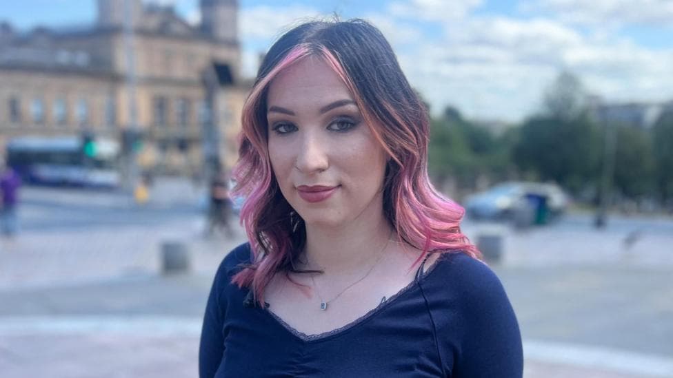 Portrait of Ella with shoulder-length dark hair featuring pink highlights, standing outdoors in a Paisley town square. She is wearing a dark long-sleeved top and a small pendant necklace. Buildings, a bus, trees, and street activity appear softly blurred in the background under a partly cloudy sky.