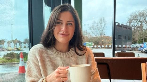 A close up image of Erin Atkinson wearing a cream jumper and holding a mug. She appears to be sitting in a public space with a car park seen out the window behind her.