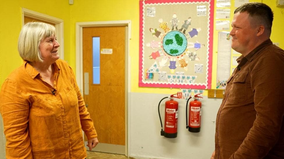 A white woman with short blonde hair with a dark yellow shirt is talking to a white man with brown hair and a brown shirt in a school corridor with drawings in the background on the wall
