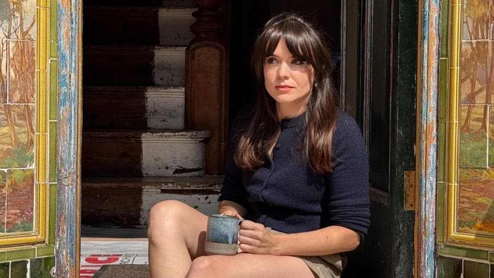 Sheri Ingram is photographed sat down in the door way of her home, which is in the process of being renovated. She is holding a blue mug which is placed on her right knee. She has long brown hair past her shoulders and is wearing a blue knitted cardigan. Steps inside her home leading upstairs can be seen behind her through the open doorway. There is Edwardian tiles on each side of the doorway, framing the photo. She is looking away from the camera and is not smiling.