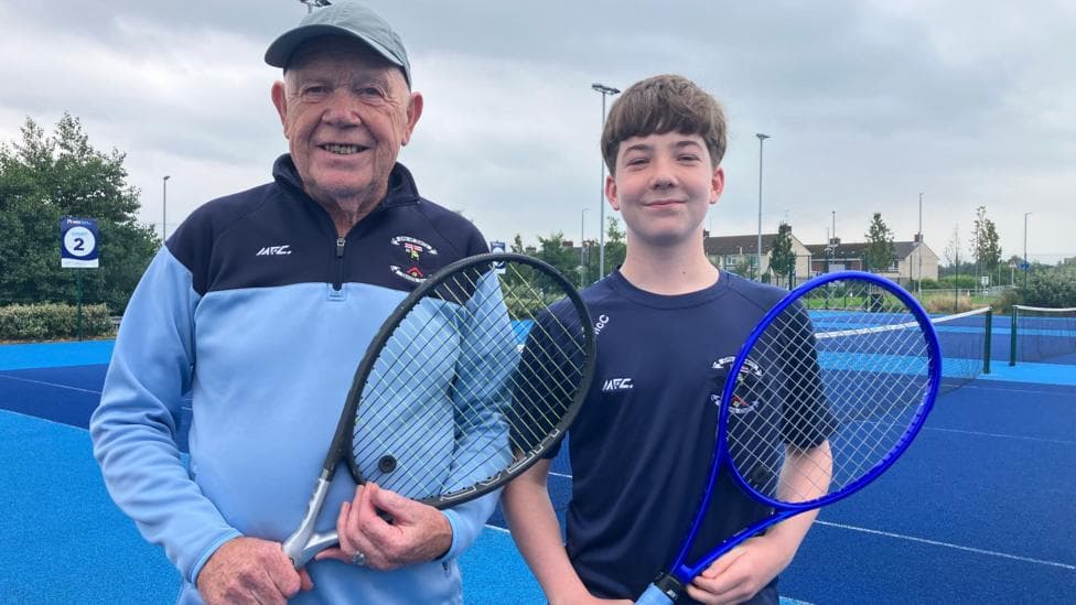 A man wearing a grey hat and blue and navy 1/4 zip and teen boy with brown hair wearing a navy top. They are both holding tennis rackets