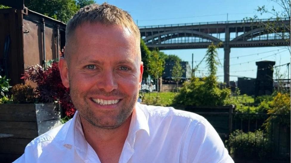 Matthew Blades smiling into the camera. His head and shoulders are visible and he has light brown short hair which is longer on the top. He has short, dark stubble around his mouth and is smiling with his death. He is wearing a white shirt which is open at the top. He is sitting in a garden in front of the High Level Bridge in Newcastle which is a tall, brown structure with arches along the side of it. There are lots of green plants and grass but on the left there is a brown shipping container. There are black fairy lights strung up above the garden and the sky above is blue.