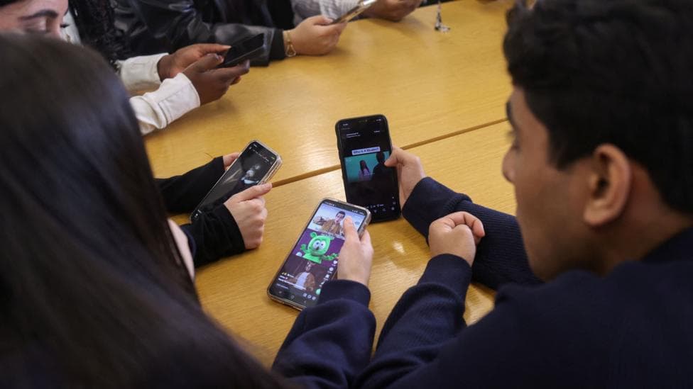 Children hold mobile phones sitting at a classroom table.