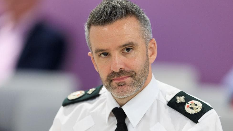 Bobby Singleton has short dark and grey hair. He is wearing a police uniform which consists of a white shirt, black tie and black badged lapels.