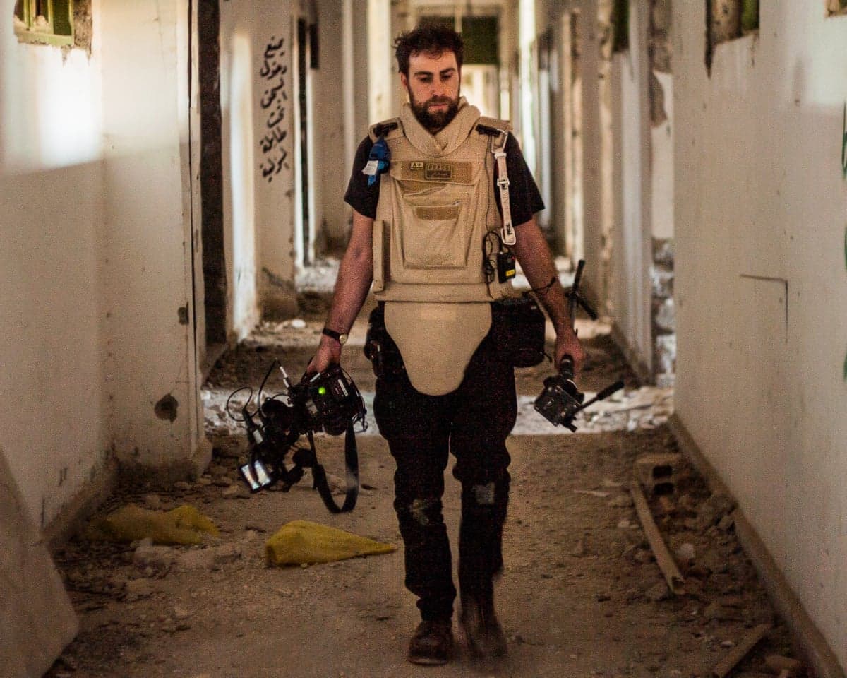 Wearing a bulletproof vest and carrying filming equipment, Josh Baker walks down a war-torn corridor