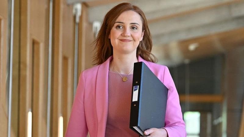Education Secretary Màiri McAllan walking down the corridor at Holyrood. She is wearing a pink blazer and top and is carrying a black ring binder folder.
