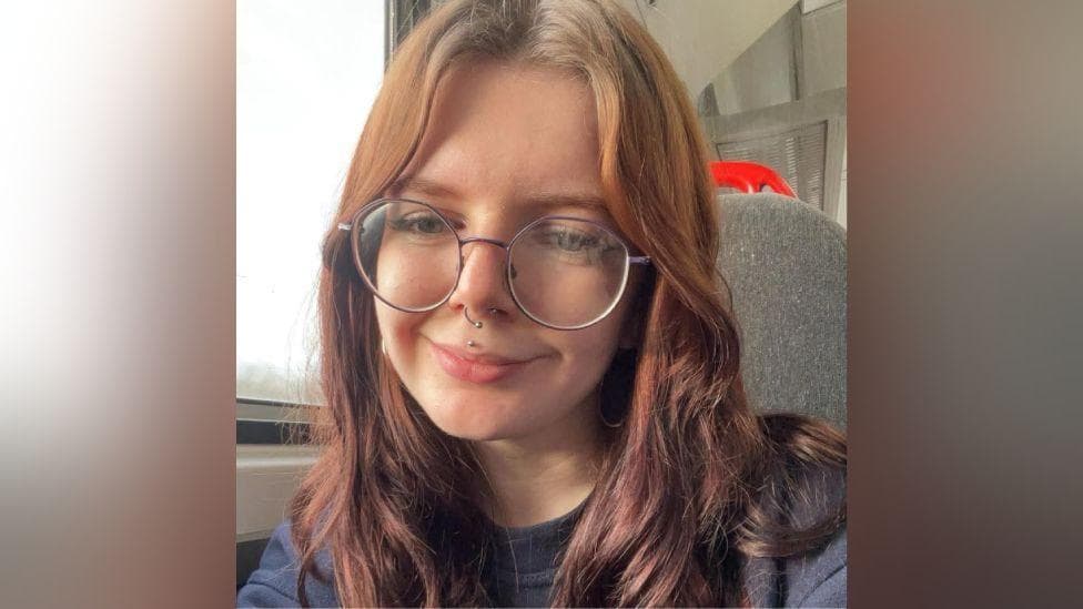 Corinna Baker. She has red hair, is smiling and is wearing a pair of large circular spectacles. She has a nose ring and stud through her lip