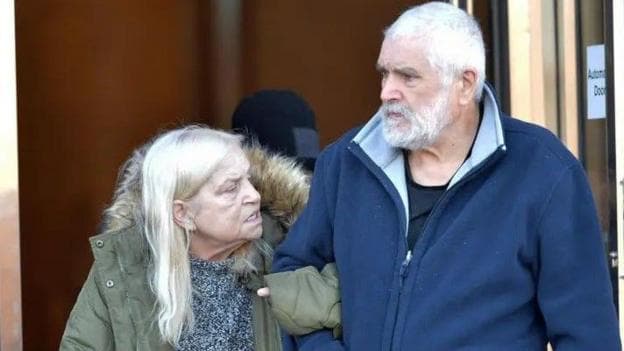 An elderly couple standing outside a courthouse, looking towards each other as they leave. The woman has long hair and is wearing a green anorak, the man has white hair, a beard and is wearing a blue fleece.