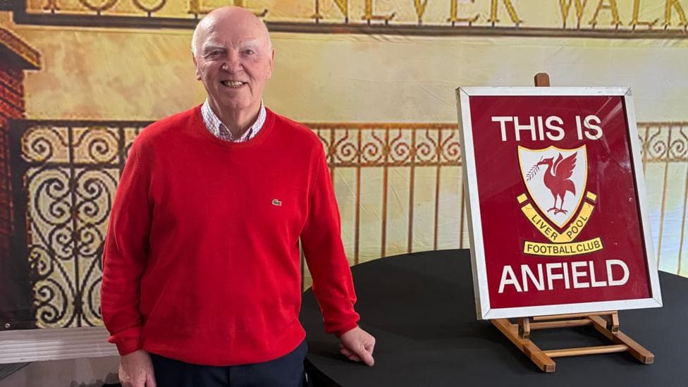A man in a red jumper stands next to the 'THIS IS ANFIELD' sign which sits on a balck table.