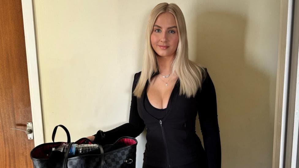 A woman stood in a hotel room with her hand on her bag