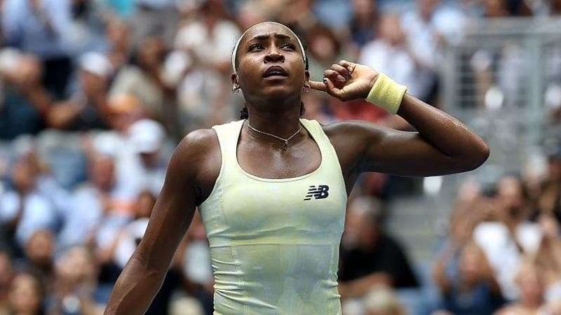 Coco Gauff, wearing a pale yellow vest, puts her left index finger behind her ear
