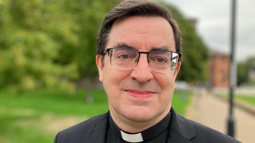 Rev Bernard Randall, a man with brown hair and glasses, wearing a black jacket and shirt, with a white dog collar.