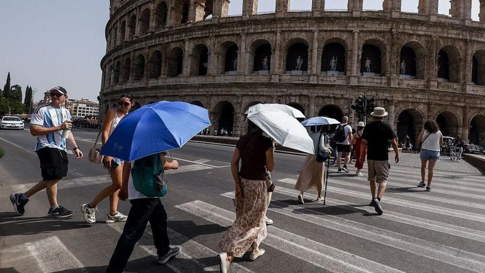A number of people are crossing the street in Rome, in the shadow of the Colosseum, - several are holding umbrellas and sunhats as protection from the Sun. The sky is blue and cloudless on the left of the picture some city buildings and trees can be seen.