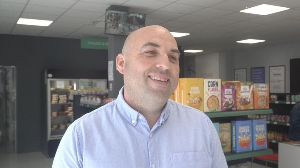 Stephen Brindley smiling and looking to the side of the frame. Supermarket shelves and fridges with goods including tins, cereal, and frozen meals are visible.