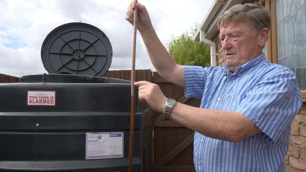 A man with light brown hair wearing a short-sleeved blue and white striped shirt stands outside by a black heating oil tank with its round lid open. In his hand he is holding a dipstick and checking the level of oil in the tank. A sign on the tank reads: This tank is alarmed.