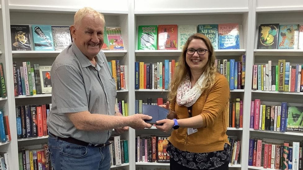 Peter meeting a librarian at Llanrwst library, handing over the late books. Both are smiling and standing in front of a row of books on shelves