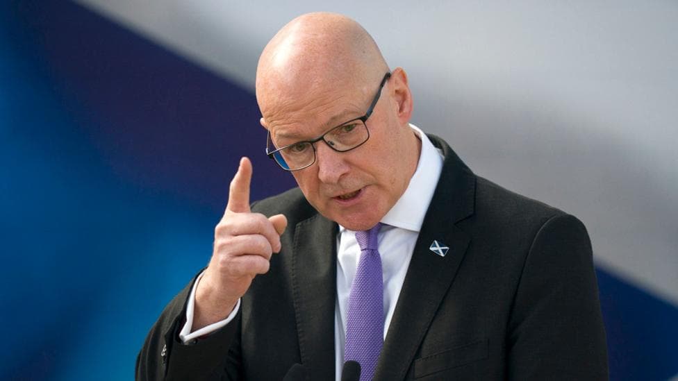 A bald headed man in glasses with a saltire badge waves his finger in the air while giving a speech