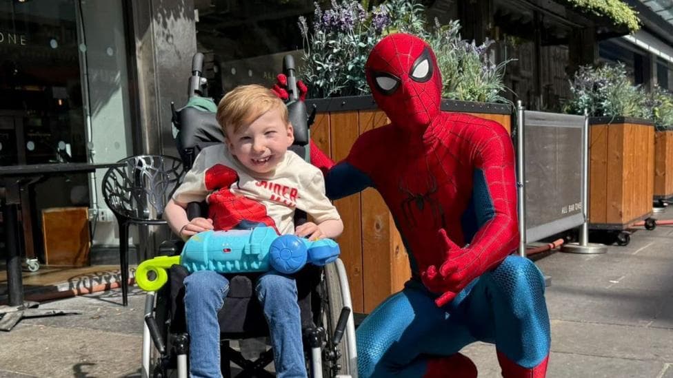 Noah Connell, who has short, blond hair, is in his wheelchair with a stunt man dressed as Spider-Man, complete with mask, posing next to him. Noah is wearing a Spider-Man T-shirt and blue jeans and is smiling