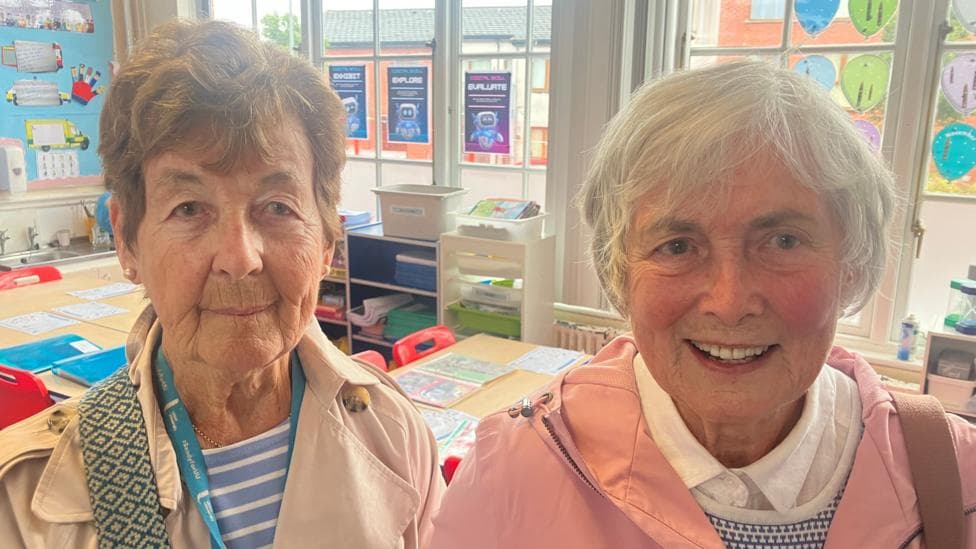 Barbara has short brown hair and is wearing a trench coat with a blue striped top. Rosaleen has short grey hair and is wearing a pink coat, white shirt and patterned vest.