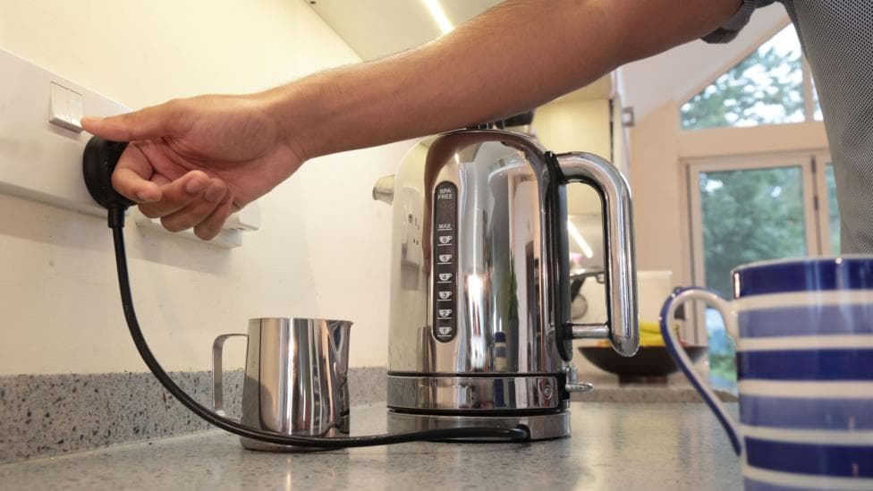 A householder switching on a domestic electricity socket to turn on a silver kettle on a kitchen counter. A silver milk jug and a blue striped mug are also on the counter.