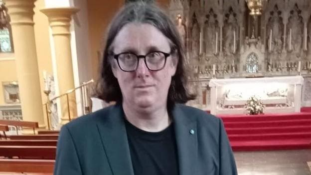 Ciaran Flanagan is wearing a grey suit and black T-shirt. He is standing in a church. He has shoulder length dark hair and is wearing black-rimmed glasses.