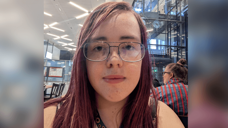 A selfie of a young woman with dyed red straight hair and glasses. She has a green sunflower lanyard round her neck.