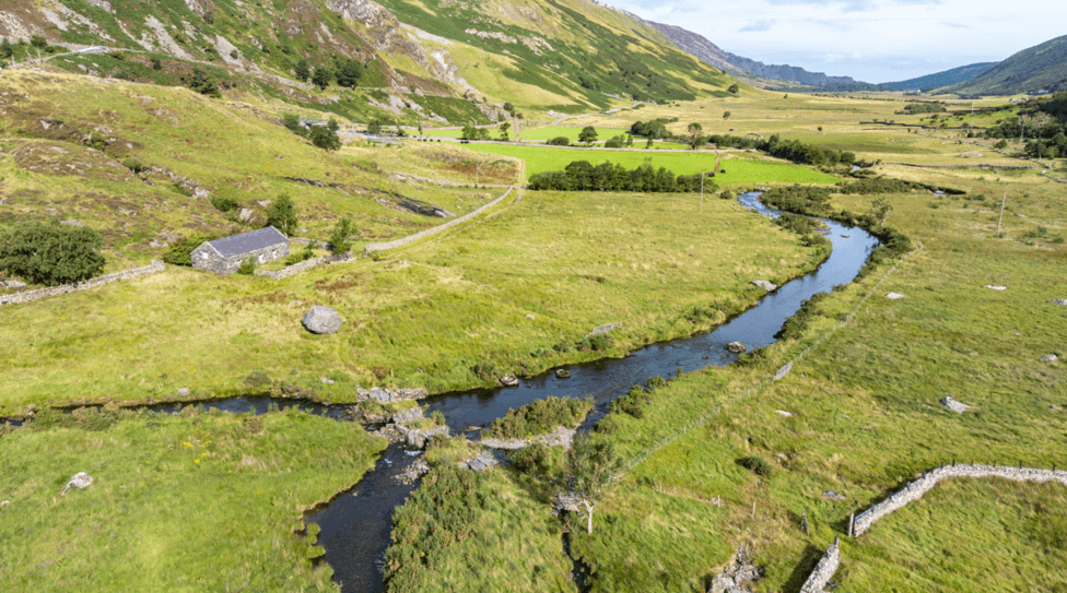 A green valley in Eryri national park. It has steep sides and is dotted with trees, a winding stream and a small stone cottage