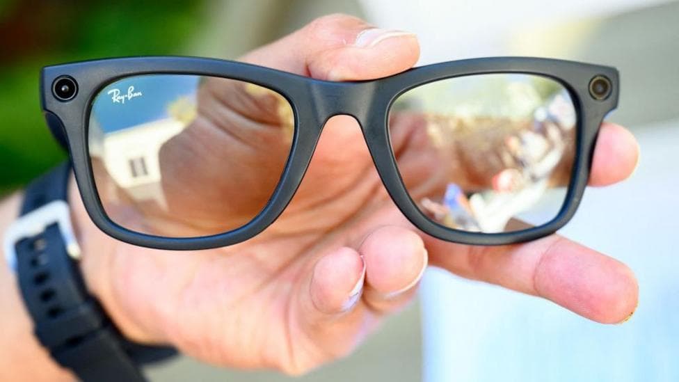 A pair of Meta glasses, being held in someone's left hand. They are black with a camera visible on each side. The person is wearing a watch.