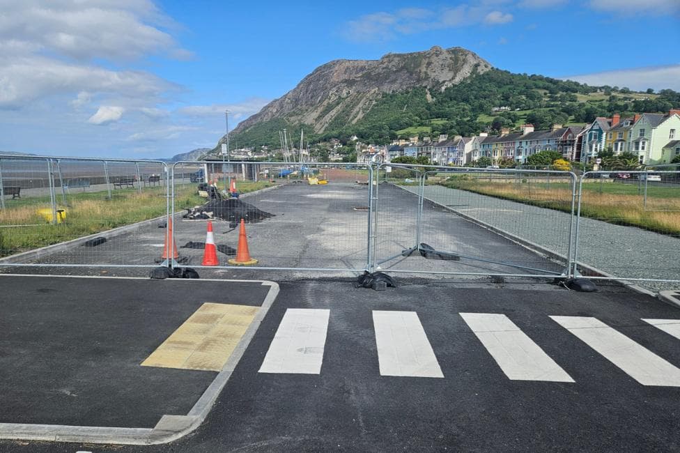 Llanfairfechan promenade car park, a metal barricade goes across a newly tarmacked area. Houses are visible lining a road in the background and there are green grass on both sides.