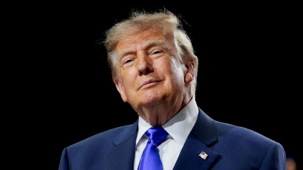 Donald Trump is wearing a navy suit jacket with a white shirt and a bright blue tie. He is smiling slightly and is standing in front of a black background.