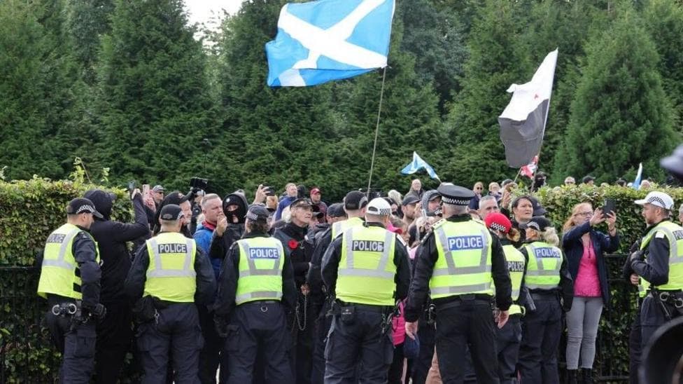 Police stand between groups at a public demonstration, one of the crowd is holding a saltire flag up in the air.