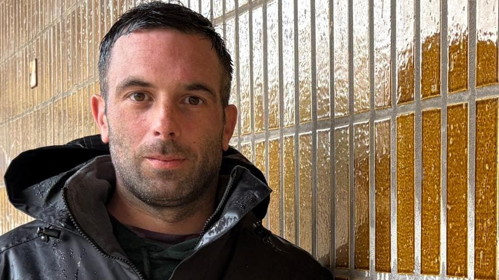 Bill Lewis looking directly to camera. He has short black hair and stubble. He is standing outside and is wearing a black hooded jacket, with rain drops over his shoulders and chest. He is leaning against a wall that is covered in rectangular tiles.