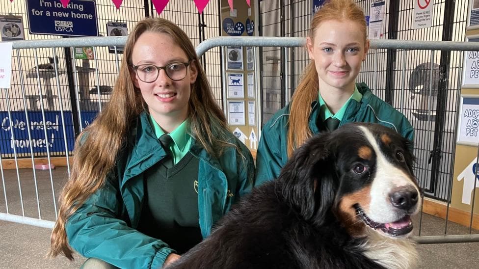 Two girls, one with dark hair and one with light hair kneel down beside a dog for a photo