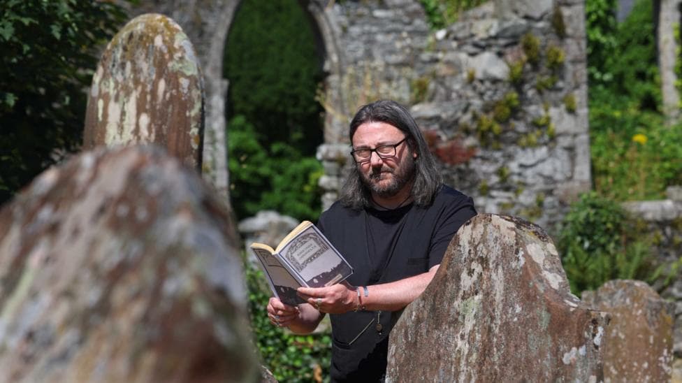 A man with long, dark hair and glasses is reading a book in an old graveyard