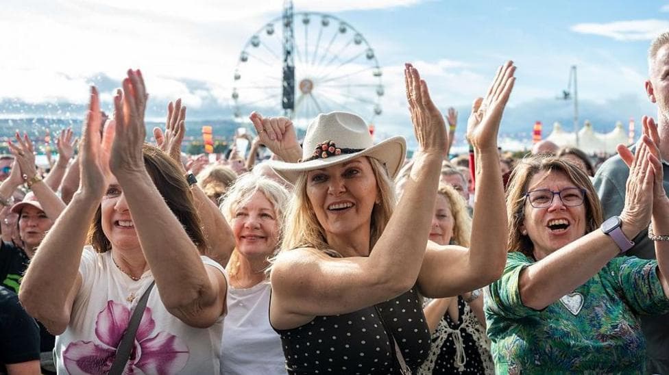 A crowd at a festival, Three women at the front are smiling while clapping in the air. The woman in the middle is wearing a stetson and a spotty top. A woman on the left is wearing a white T-shirt with a large pink floral design and a woman on the right is wearing blue-framed glasses, a colourful patterned top, and a smartwatch. There is a big wheel behind them.
