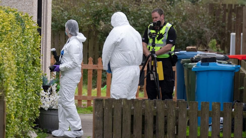 Two officers in white hazmat suits and head coverings in a garden. One of them is carrying a camera. There is also a uniformed police officer in the garden and a camera on a tripod. There are also wheelie bins and fences.