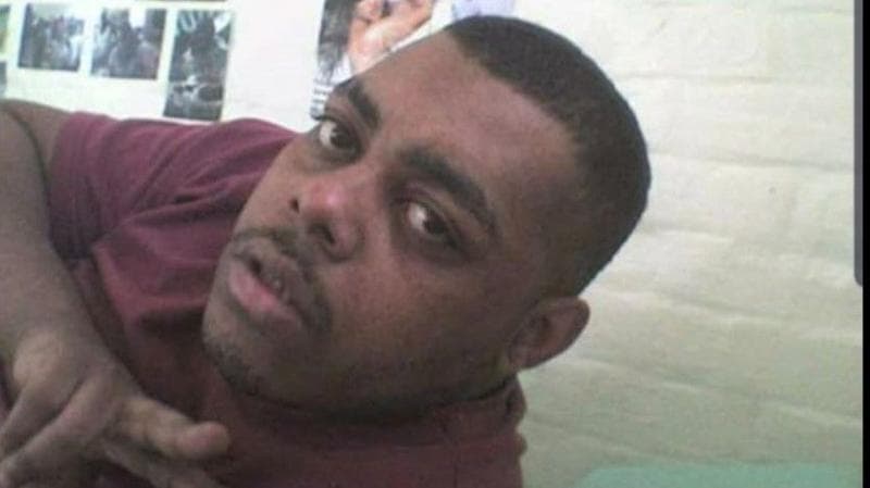 A selfie of Leroy. He is pointing at the camera, with his head and shoulders visible. He is wearing a burgundy t-shirt and has stubble and short black hair. A white brick background with posters on the wall can be seen behind him. It is a head and shoulders shot of him.