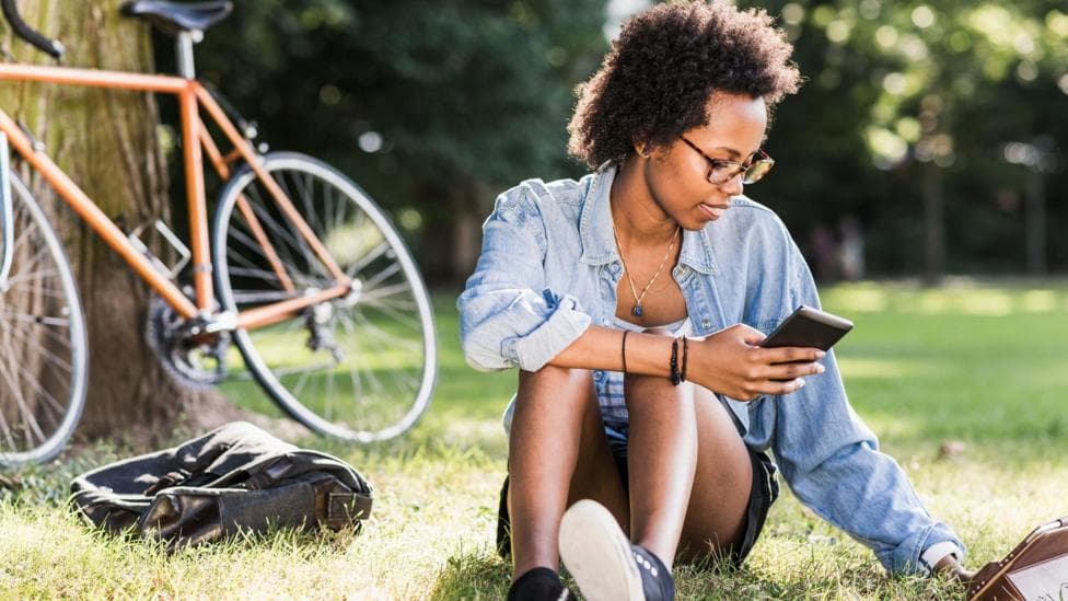 Student sits on the grass with a phone in her hand, a tablet computer on the grass and a bike resting on a tree behind her.