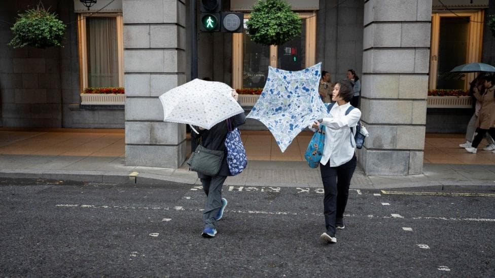 Two people cross the road with umbrellas, one has been turned inside out by the wind