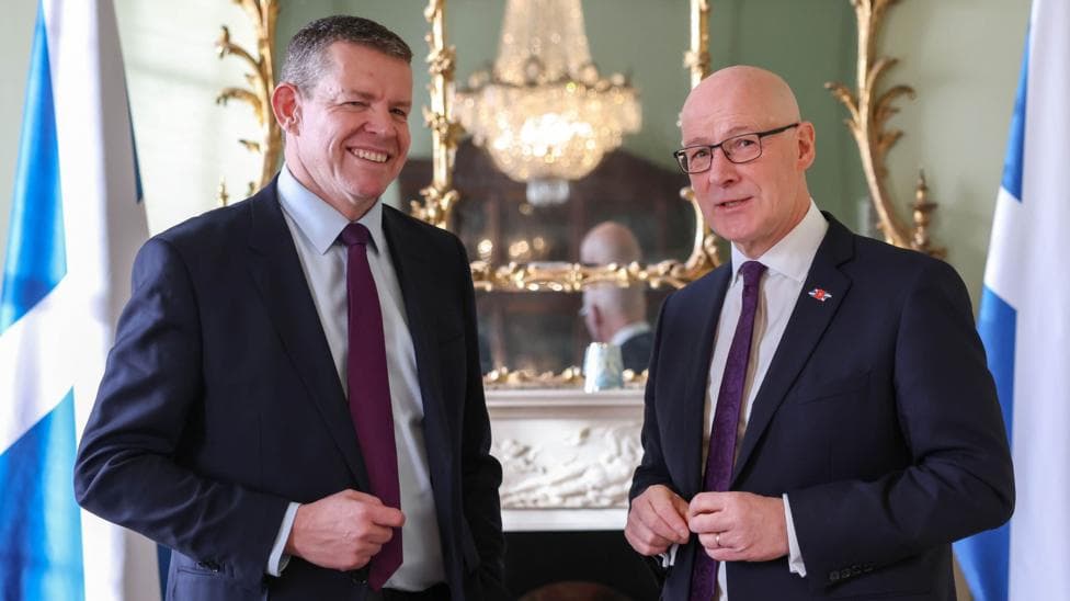 Two men in dark suits, Rhun ap Iorwerth and John Swinney, standing in front of two saltire flags and an ornate fireplace.