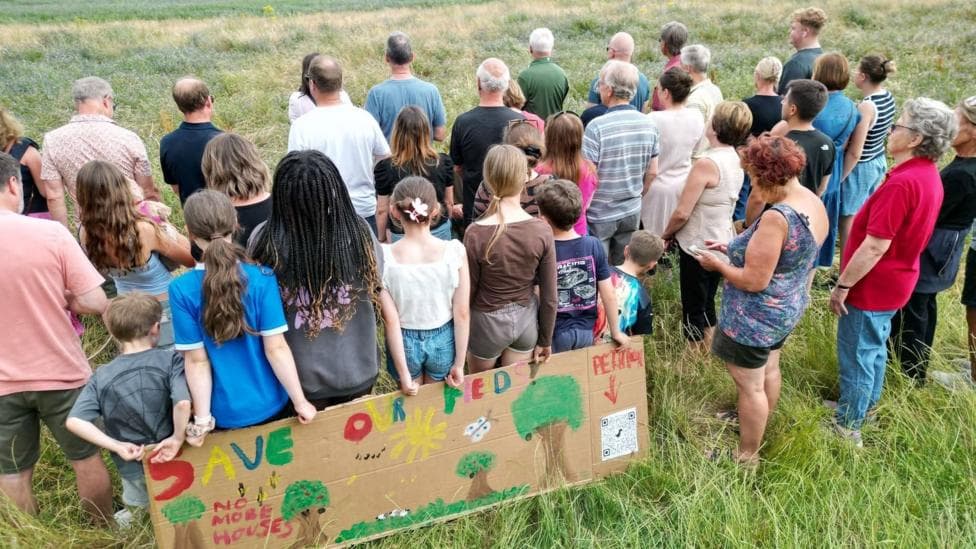 The back of a group of people stood on a field with a sign hat says save our fields