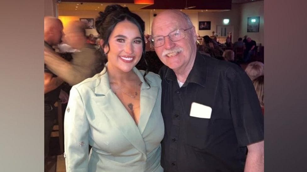 A woman with dark hair, wearing a green jacket, stands beside her grandfather. He is wearing a black shirt and black trousers. They are in a restaurant.