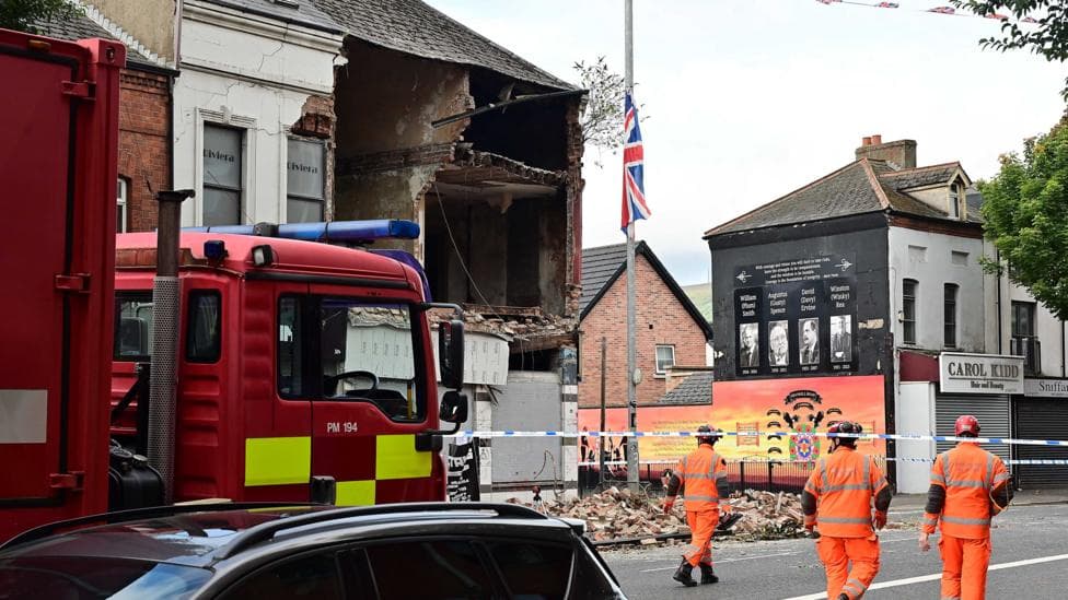 A partically collapsed building with the 3 workmen in orange suits in the foreground. There is part of a fire engine and the roof of a car visible.