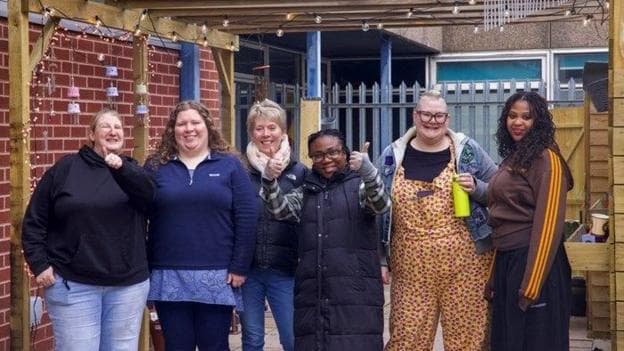A group of six woman all dressed in outdoor warm clothing are stood in a line as they smile at the camera. Two women have their thumbs up. They are stood outside under a wooen canopy