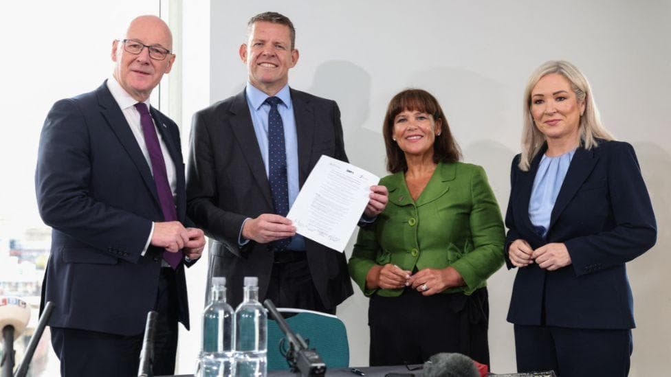 Four politicians in business wear smile while one in the centre holds a signed document