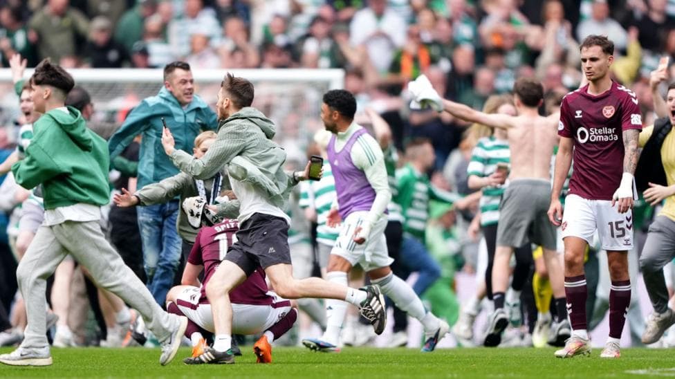 Celtic fans rush onto the pitch and wildly celebrate, many wearing replica green and white hooped jerseys. A despondent Hearts player, wearing a maroon shirt, white shorts and maroon socks, is walking off the pitch.