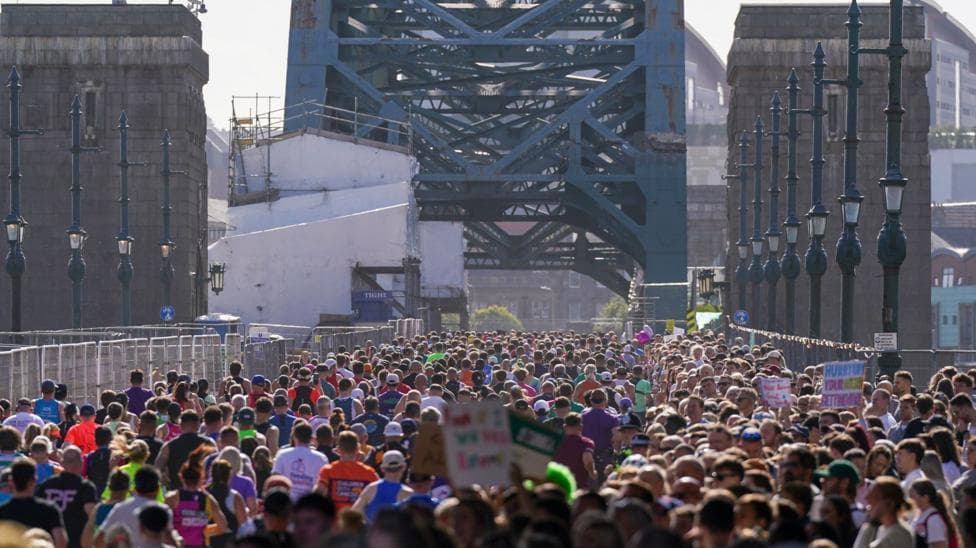 The backs of hundreds of runners running across the Tyne Bridge which is partially covered by scaffolding on the left. Spectators stand on the right, many holding up colourful signs and looking to the runners on the left.