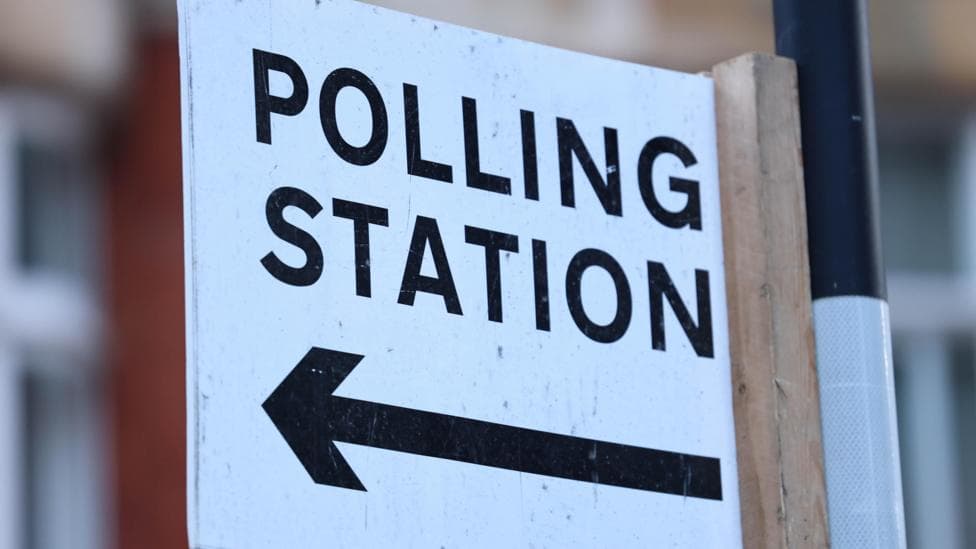 A polling station sign on a post. Polling station is written in black on white background, with an arrow pointing to the left below the words