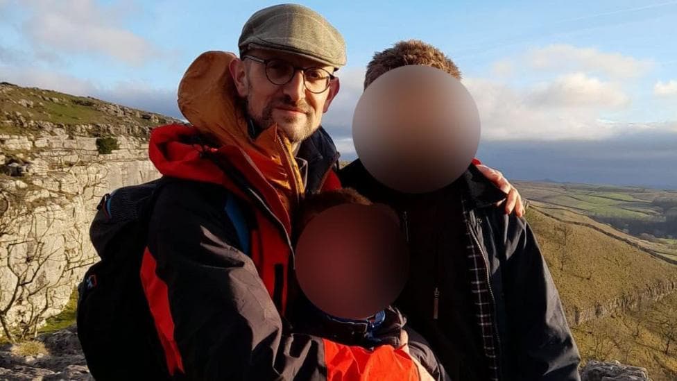 Hector Patemore wearing a red and black jacket, flat cap and glasses. He is hillwalking with two children, whose faces are blurred.