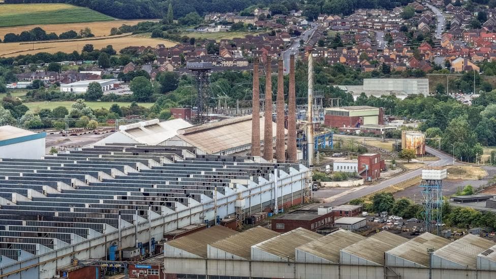 A steelworks site, pictured against the backdrop of the skyline in Rotherham. Four tall brick towers stand above the rest of the complex buildings. Residential homes can be seen in the far distance, with green fields to the left.